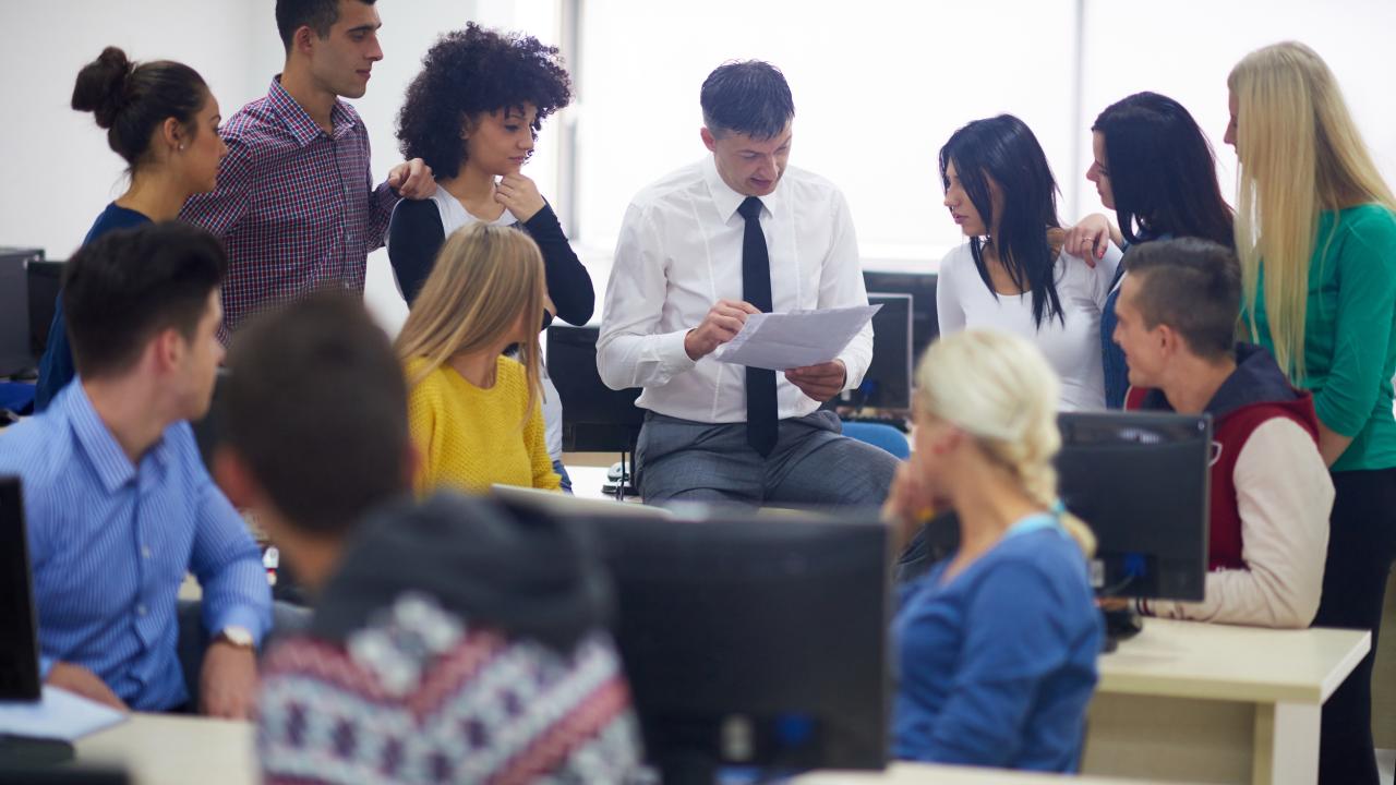 group of older students standing around, listening intently to a faculty member who wearing a white shirt and tie in a computer room. The faculty member's head is down as he's pointing to a piece of paper in his hand,